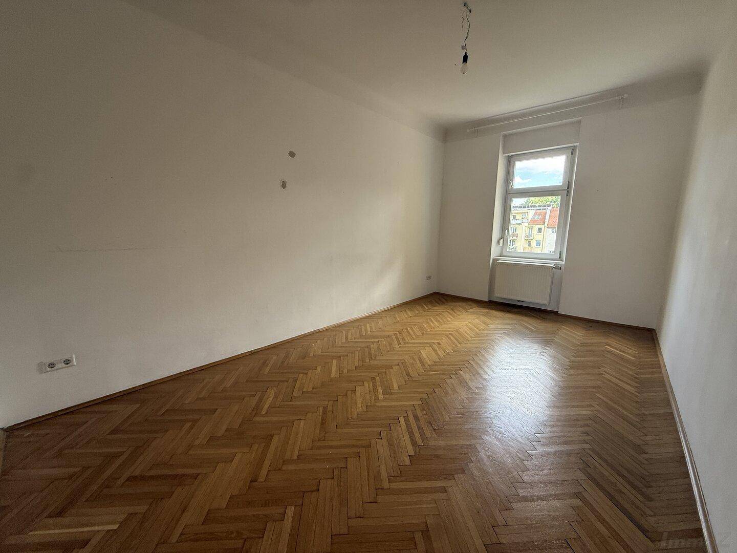 Ruhiges Zimmer mit Fischgrätparkett, weißen Wänden und einem Fenster, ideal als Schlafzimmer.