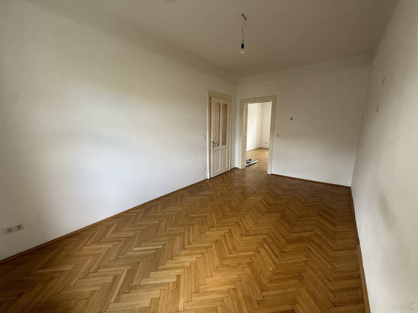 Geräumiges Zimmer mit Fischgrätparkett und weißen Wänden, das durch eine Tür verbunden ist.