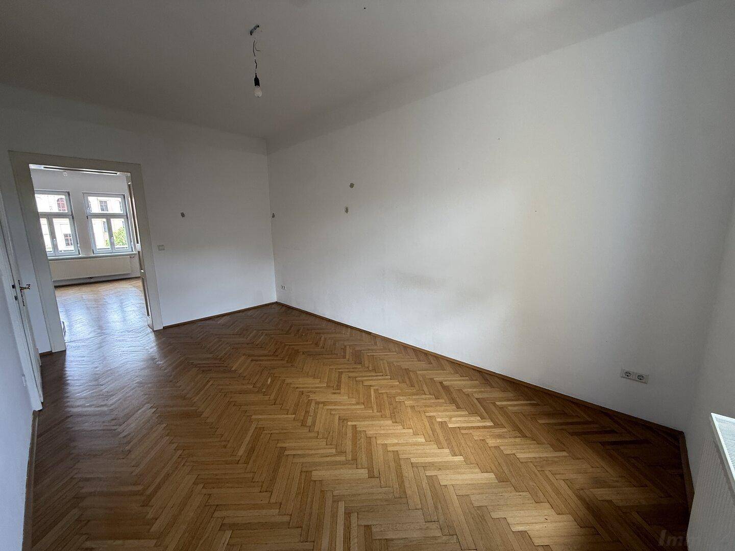Helles Zimmer mit Fischgrätparkett und offenem Durchgang zu einem weiteren Raum mit Fenstern.