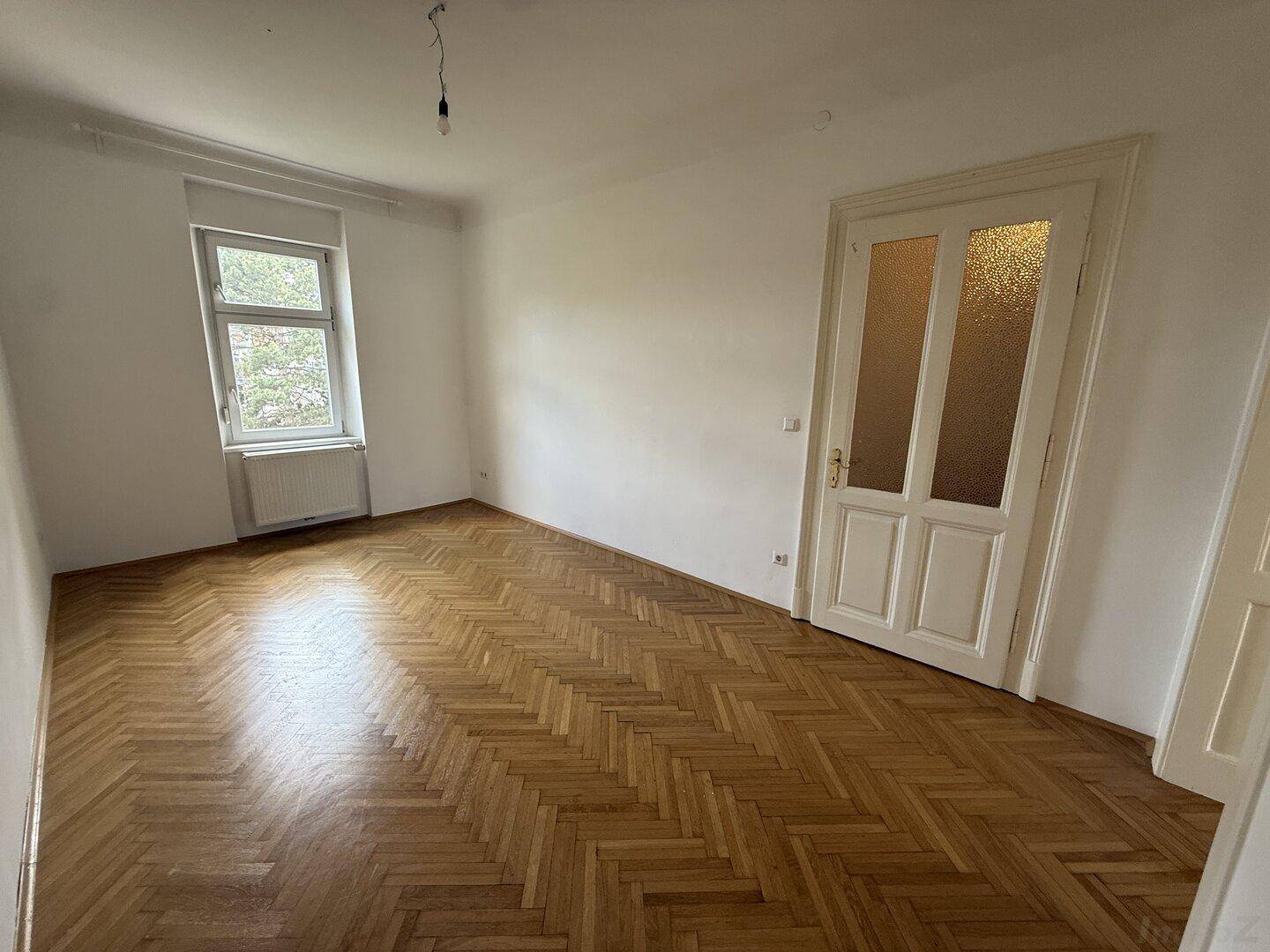 Helles Zimmer mit Fischgrätparkett, einem Fenster und einer Tür mit Milchglaseinsätzen.