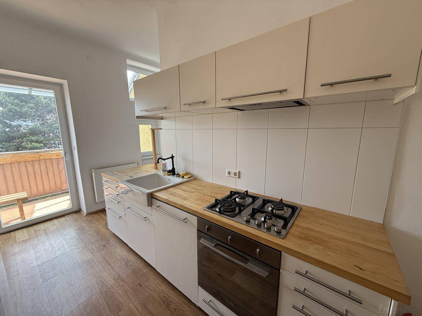 Moderne Küche mit weißen Schränken, Holz-Arbeitsplatte, Gasherd und einem Fenster mit Balkonzugang.