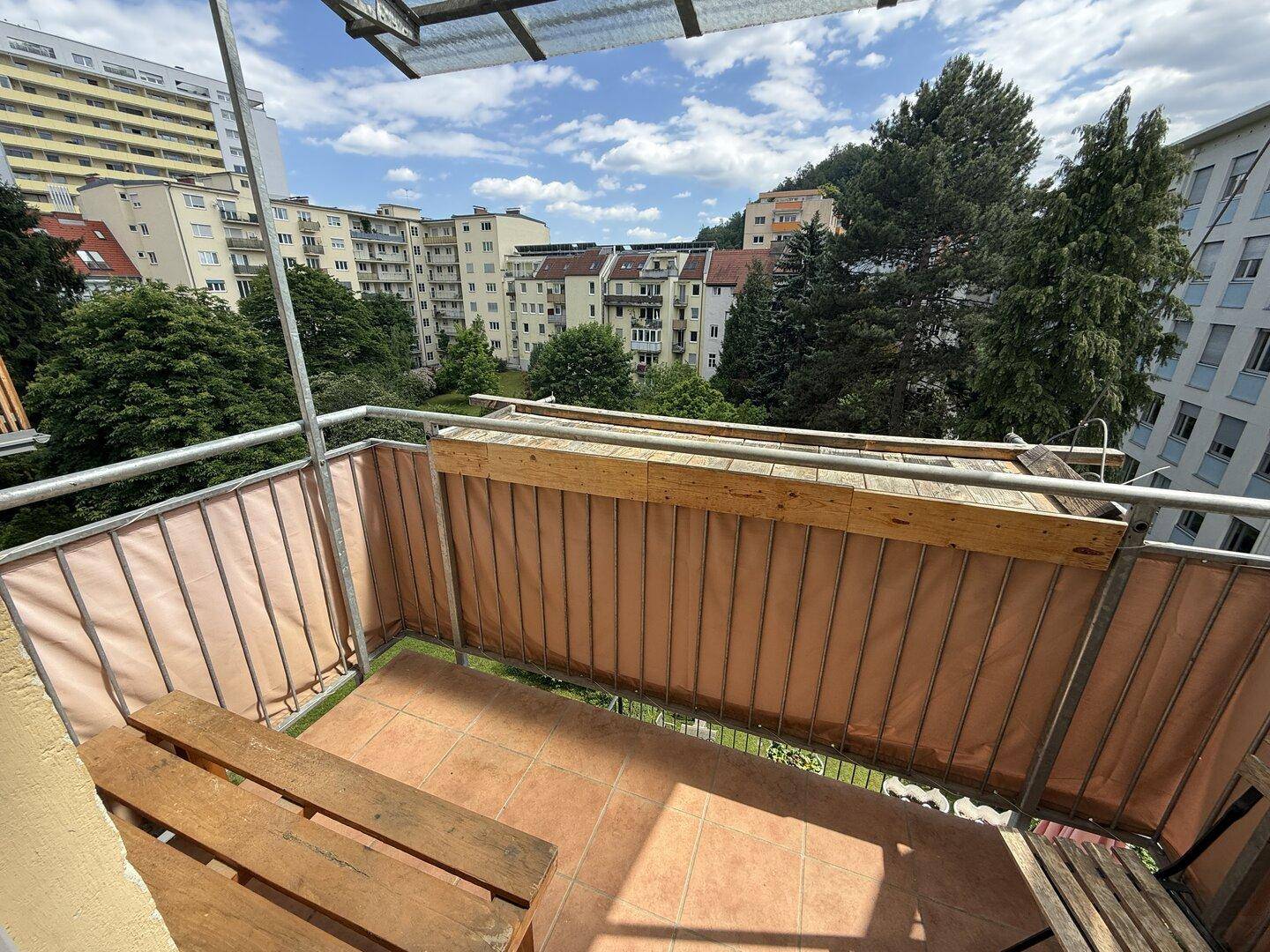 Sonniger Balkon mit Fliesenboden, Holzbank und Blick auf umliegende Wohngebäude und Bäume.