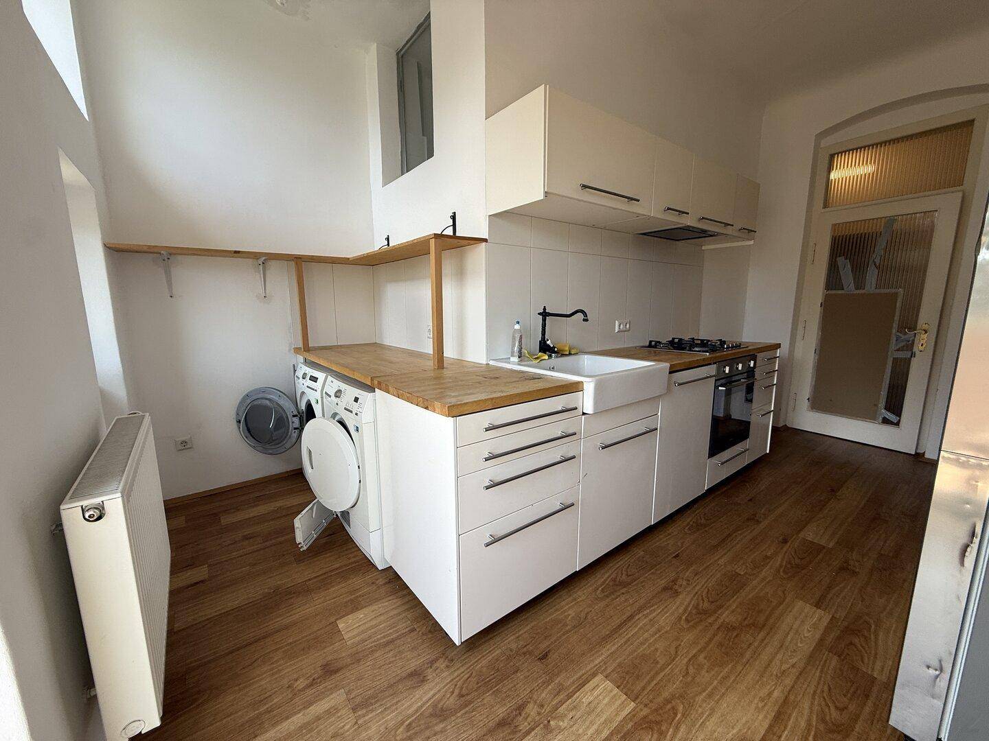 Funktionale Küche mit weißen Schränken, Holz-Arbeitsplatte und integrierter Waschmaschine unter Regalen.