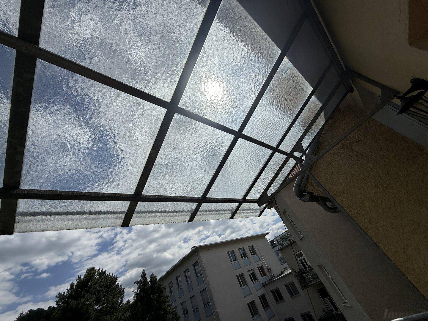 Blick vom Balkon nach oben auf eine Glasüberdachung und den blauen Himmel mit Wolken.