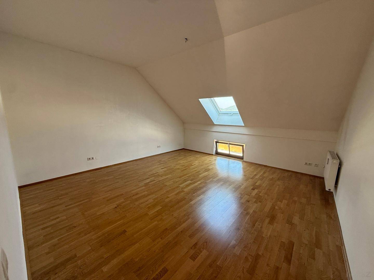 Helles Dachzimmer mit Holzboden, schrägen Wänden und einem einzelnen Dachfenster für natürliches Licht.