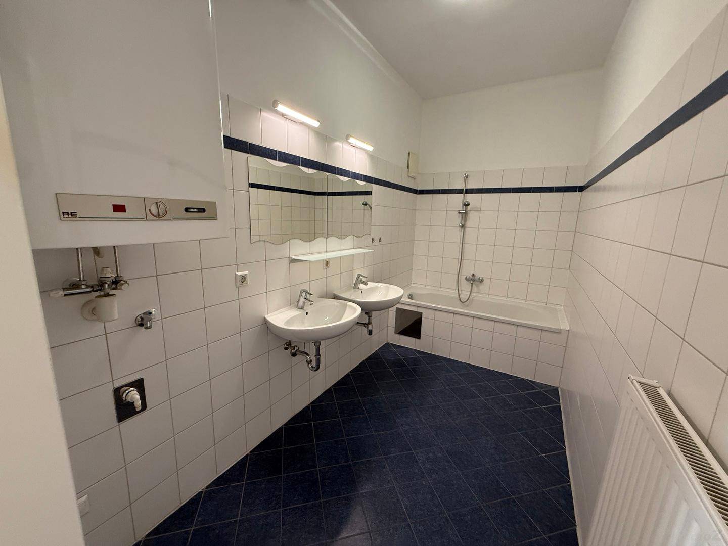 Badezimmer mit weißen Wandfliesen, blauem Fliesenboden, Doppelwaschbecken und Badewanne mit Duschfunktion.