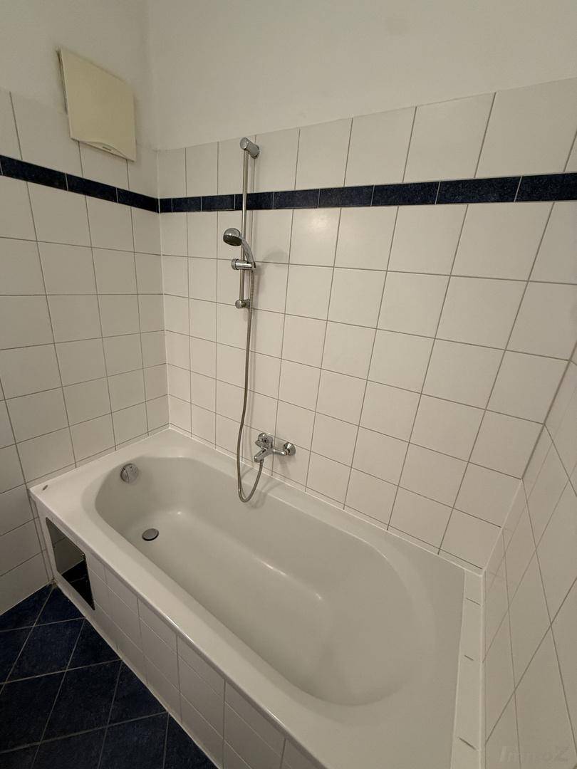 Badewanne mit Duscharmatur, umgeben von weißen Fliesen und einem blauen Fliesenboden.