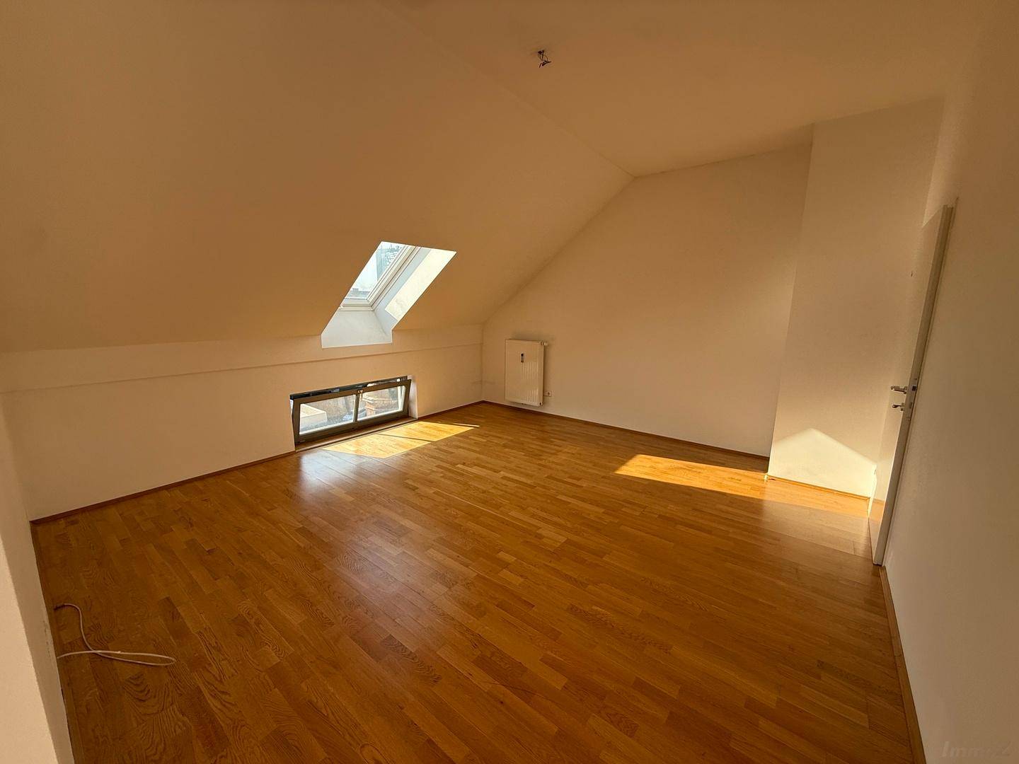 Helles Dachzimmer mit Holzboden, schrägen Wänden und einem einzelnen Dachfenster für natürliches Licht.