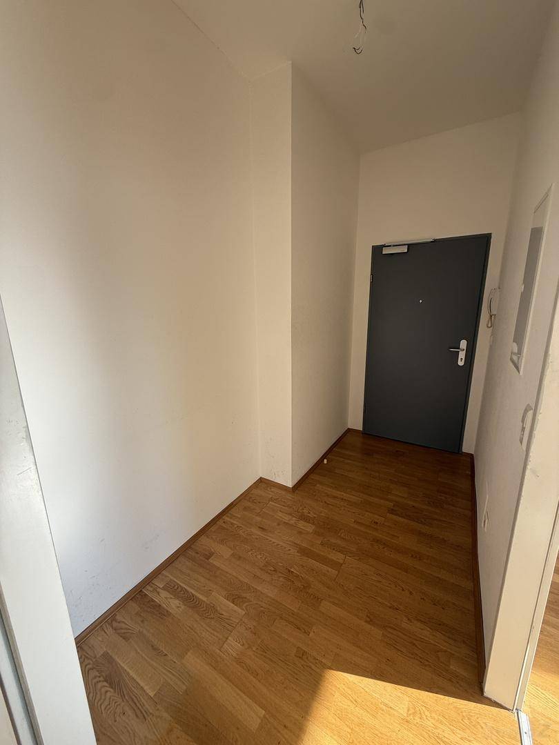 Eingangsbereich mit Holzboden, weißen Wänden und einer dunklen Eingangstür, die in die Wohnung führt.
