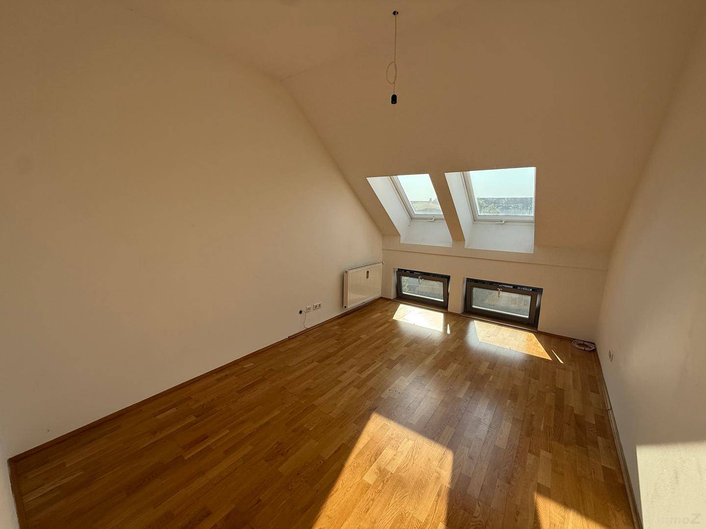 Geräumiges Dachzimmer mit Holzboden, schrägen Wänden und großen Fenstern, die viel Sonnenlicht hereinlassen.