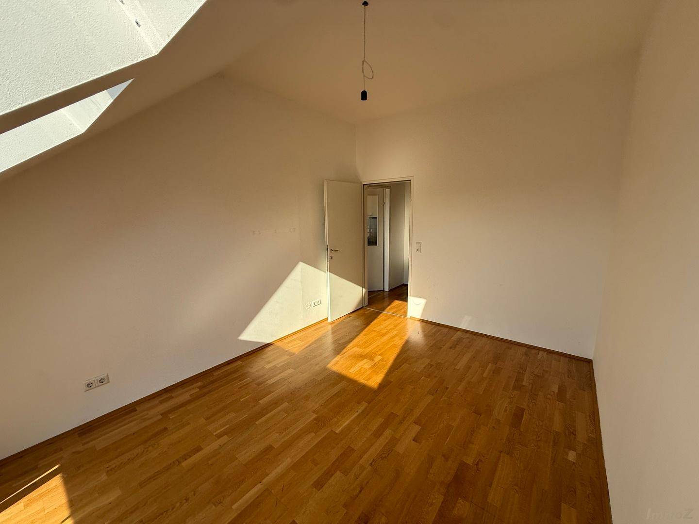Lichtdurchflutetes Dachzimmer mit Holzboden und Zugang zu einem weiteren Raum, ideal als Schlafzimmer.