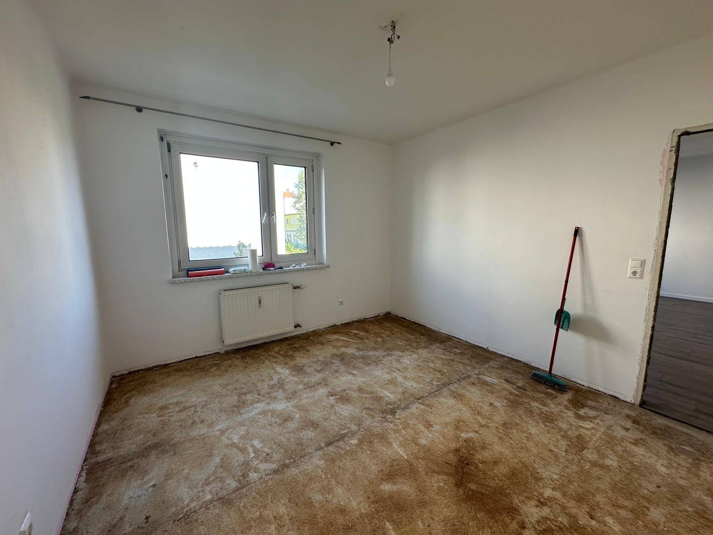 Helles Zimmer mit Fenster, vorbereitetem Bodenbelag und weißen Wänden, wartet auf neue Gestaltung.