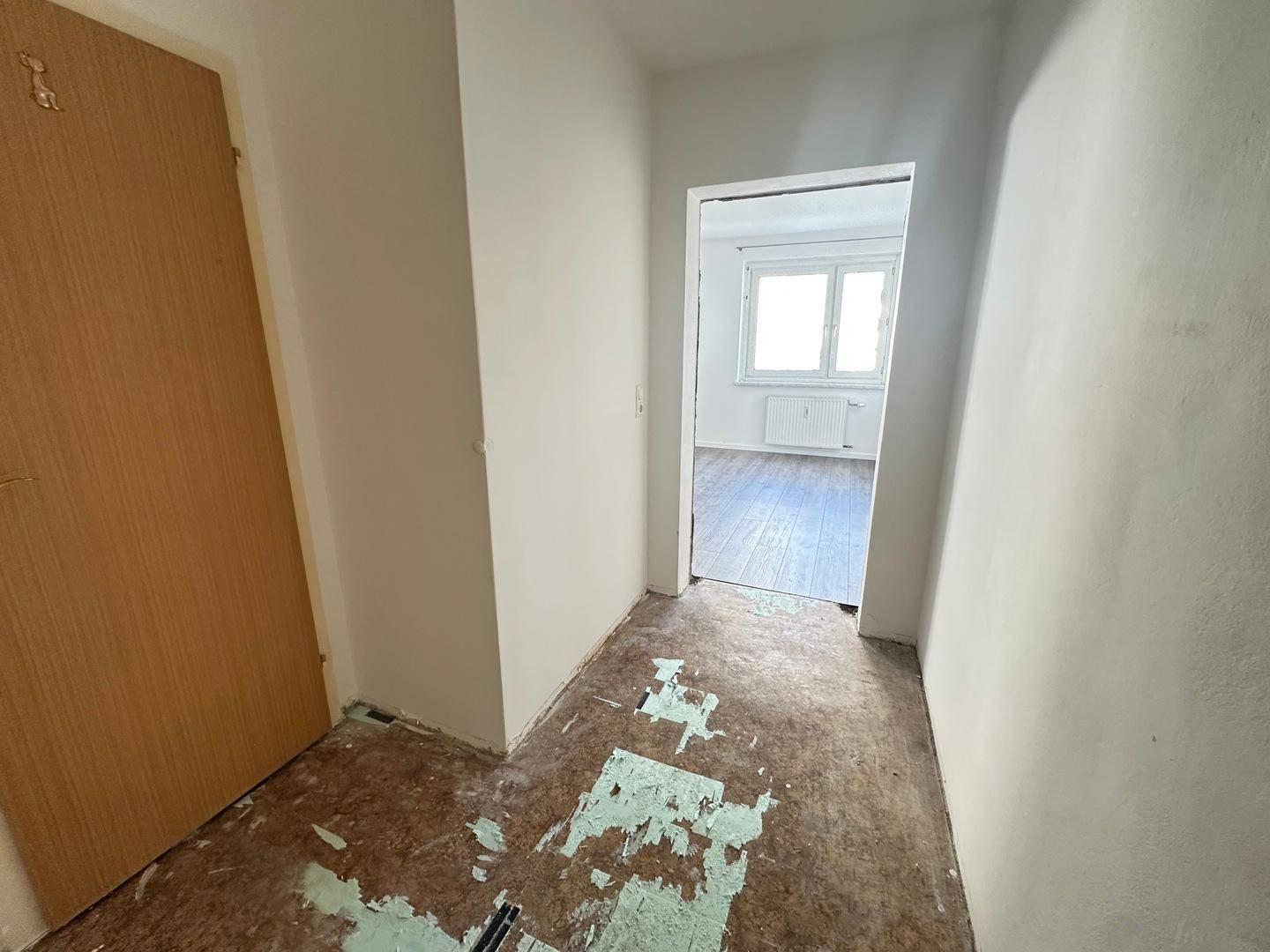 Flur mit vorbereitetem Bodenbelag und Zugang zu einem hellen Zimmer mit Laminat.