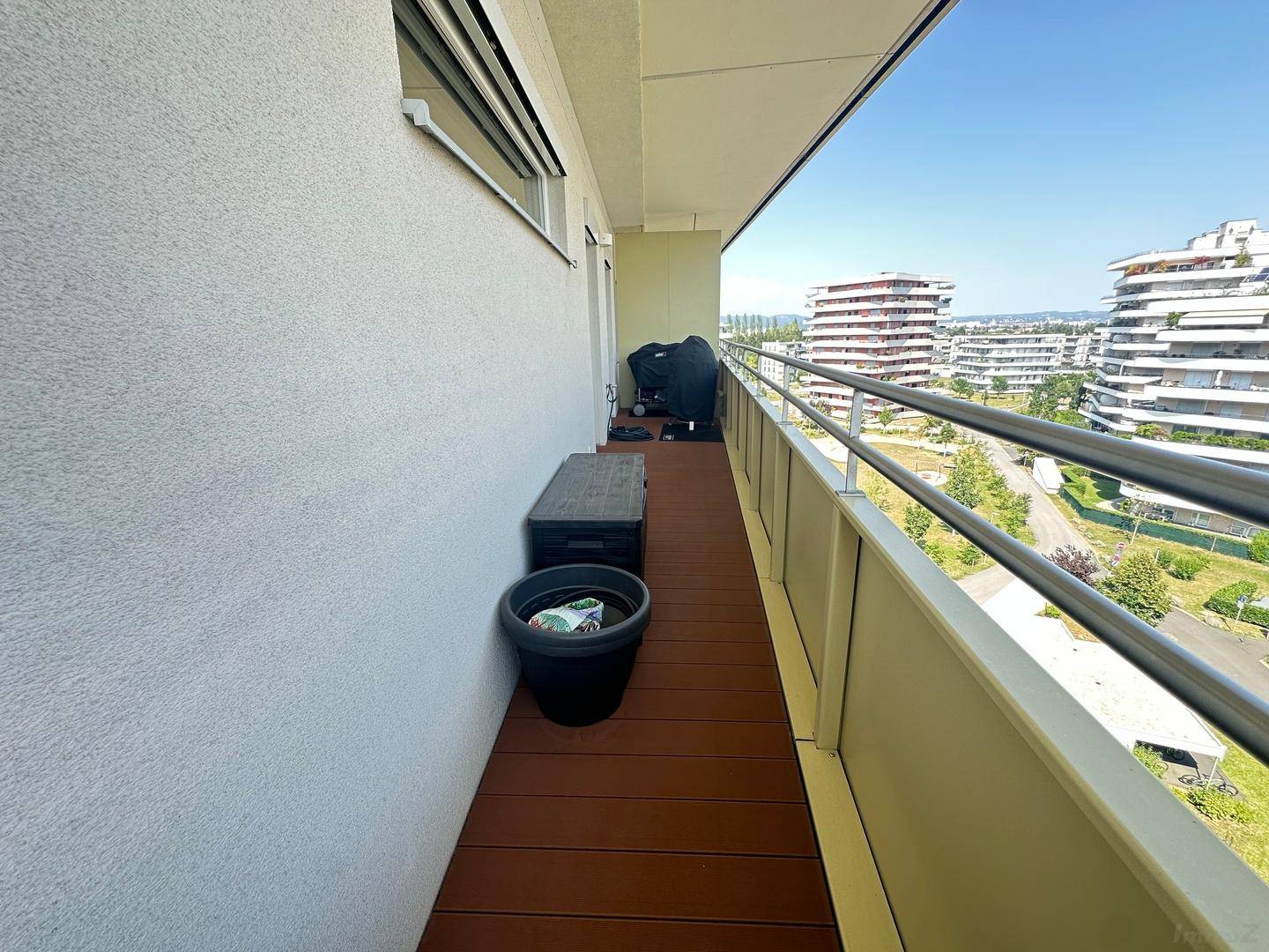 Langer Balkon mit einer Aufbewahrungsbox und Blick auf die moderne Wohnbebauung unter blauem Himmel.