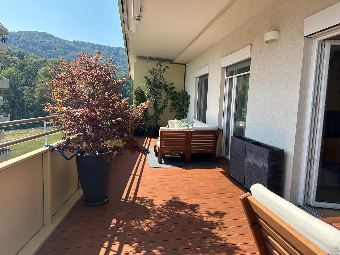 Heller Balkon mit bequemen Outdoor-Möbeln, Pflanzen und sonnigem Blick auf die umliegenden Gebäude und Berge.