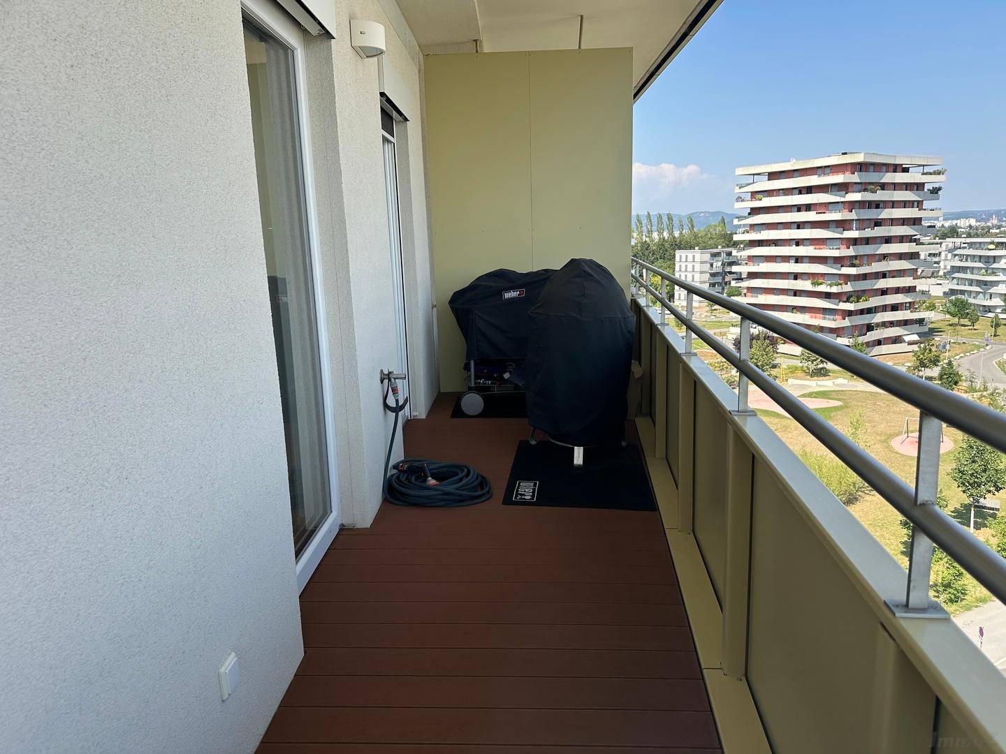 Langer Balkon mit Grillbereich, Schlauch und Blick auf moderne Wohngebäude in der Ferne.