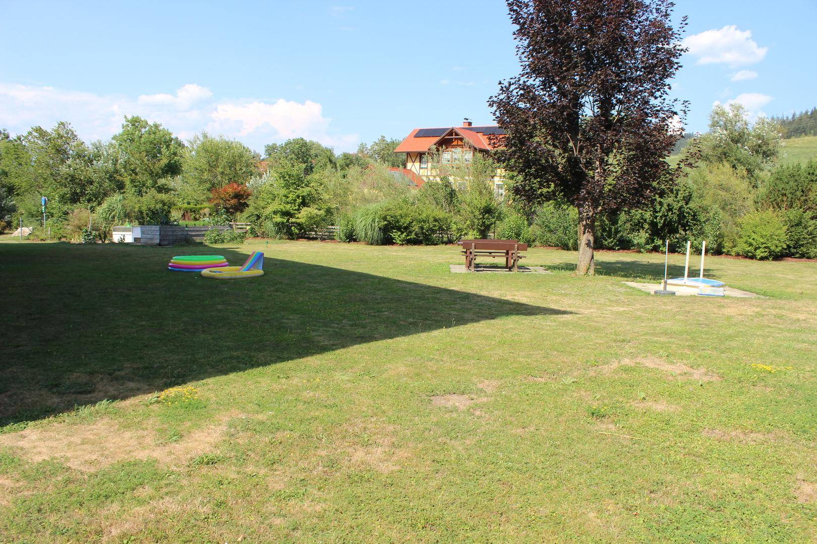 Weitläufige Rasenfläche im Gartenbereich mit Sitzbank und Spielgeräten im Hintergrund.