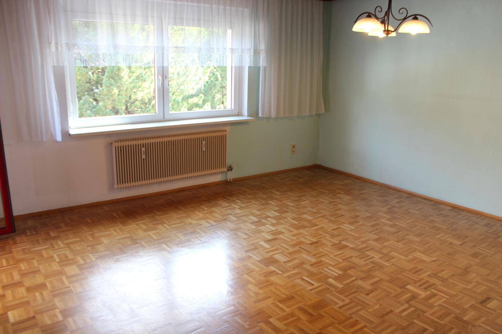 Helles Zimmer mit Parkettboden, großen Fenstern und einem Heizkörper unter dem Fenster.