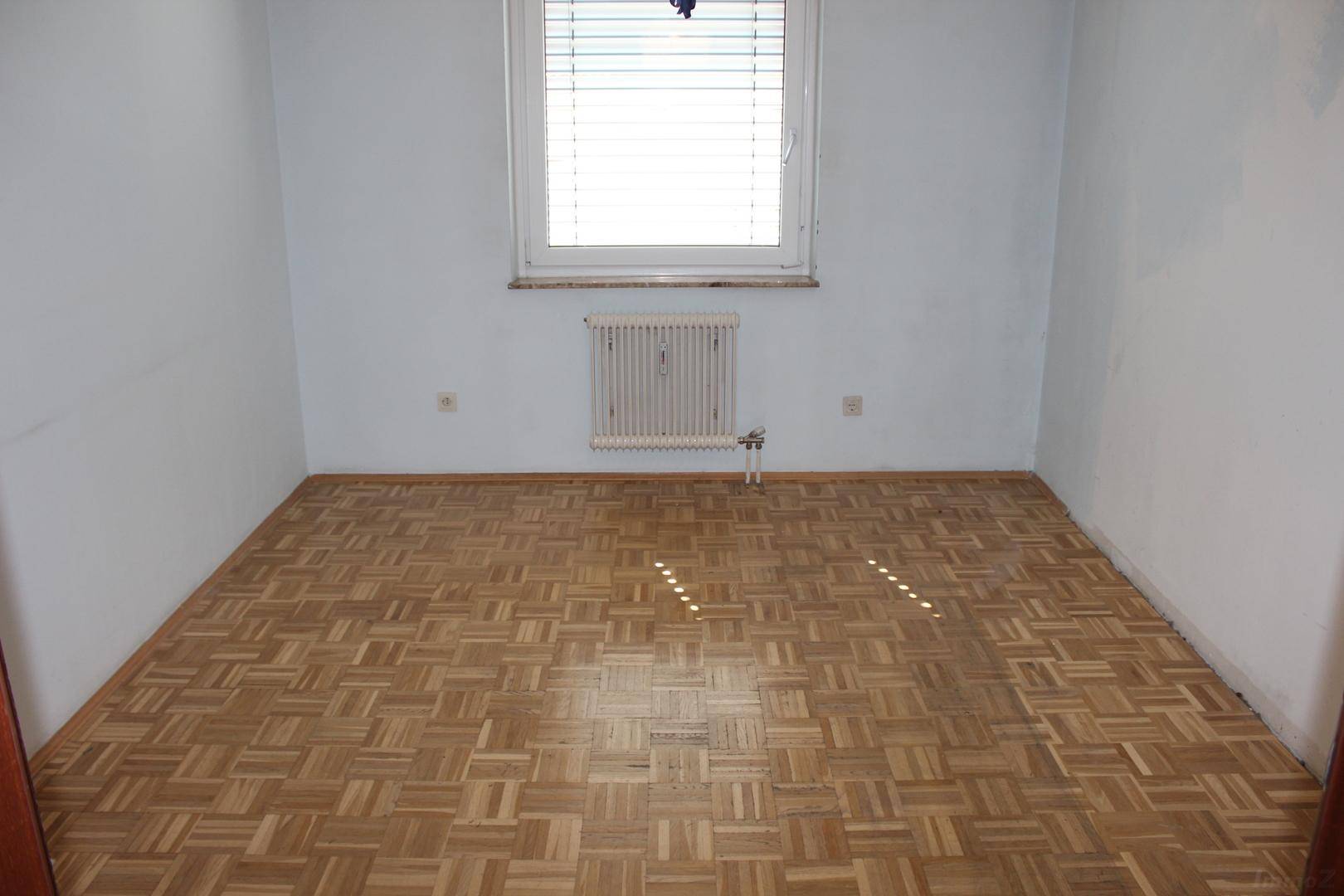 Schlafzimmer mit Parkettboden, einem Fenster mit Jalousien und einem Heizkörper.