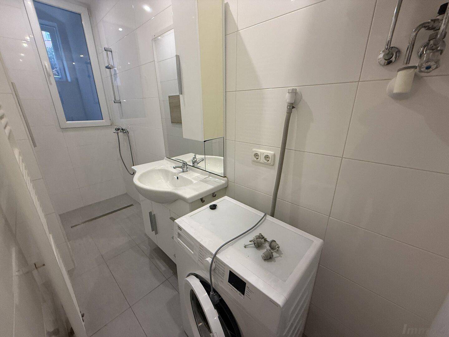 Modernes Badezimmer mit weißen Fliesen, Waschbecken, Spiegelschrank und integrierter Waschmaschine.