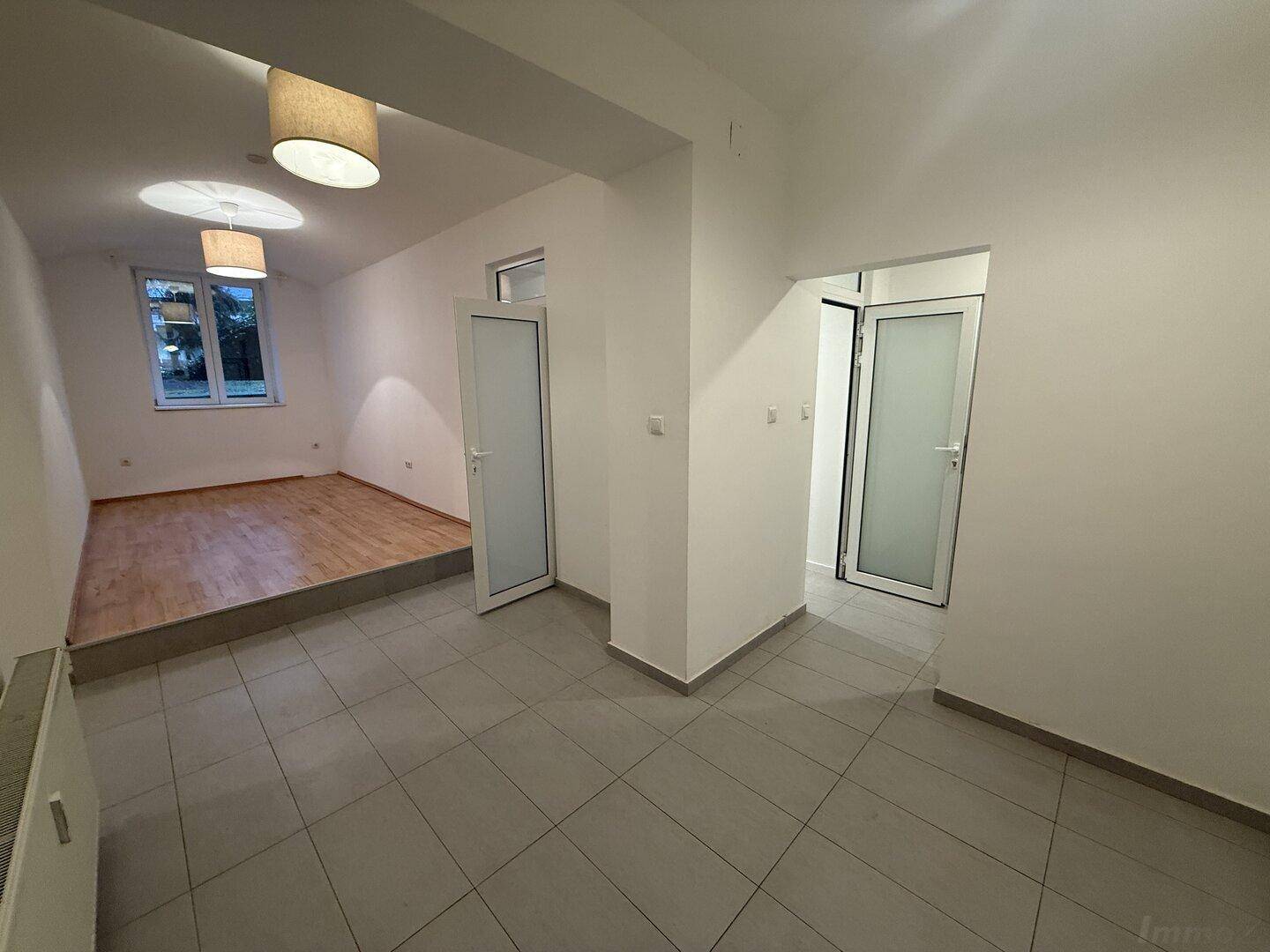 Blick vom gefliesten Bereich in ein erhöhtes Zimmer mit Holzfußboden und Fenster.