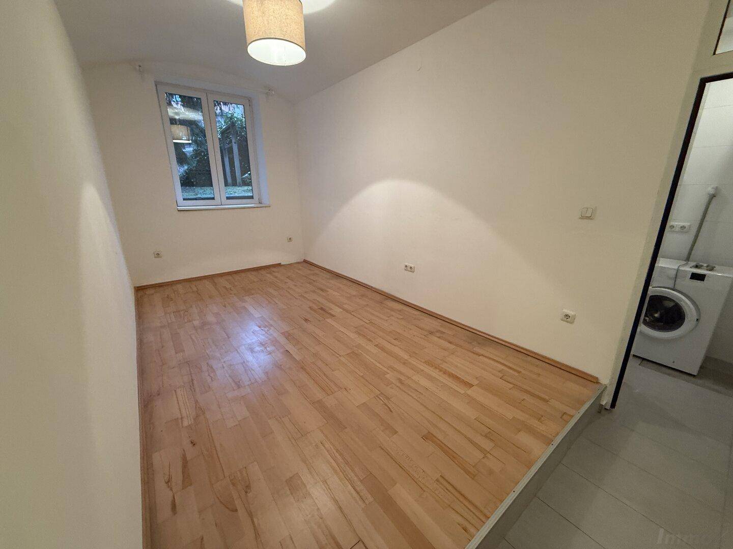 Helles Zimmer mit Holzfußboden und Fenster, angrenzend an einen Bereich mit Waschmaschine.