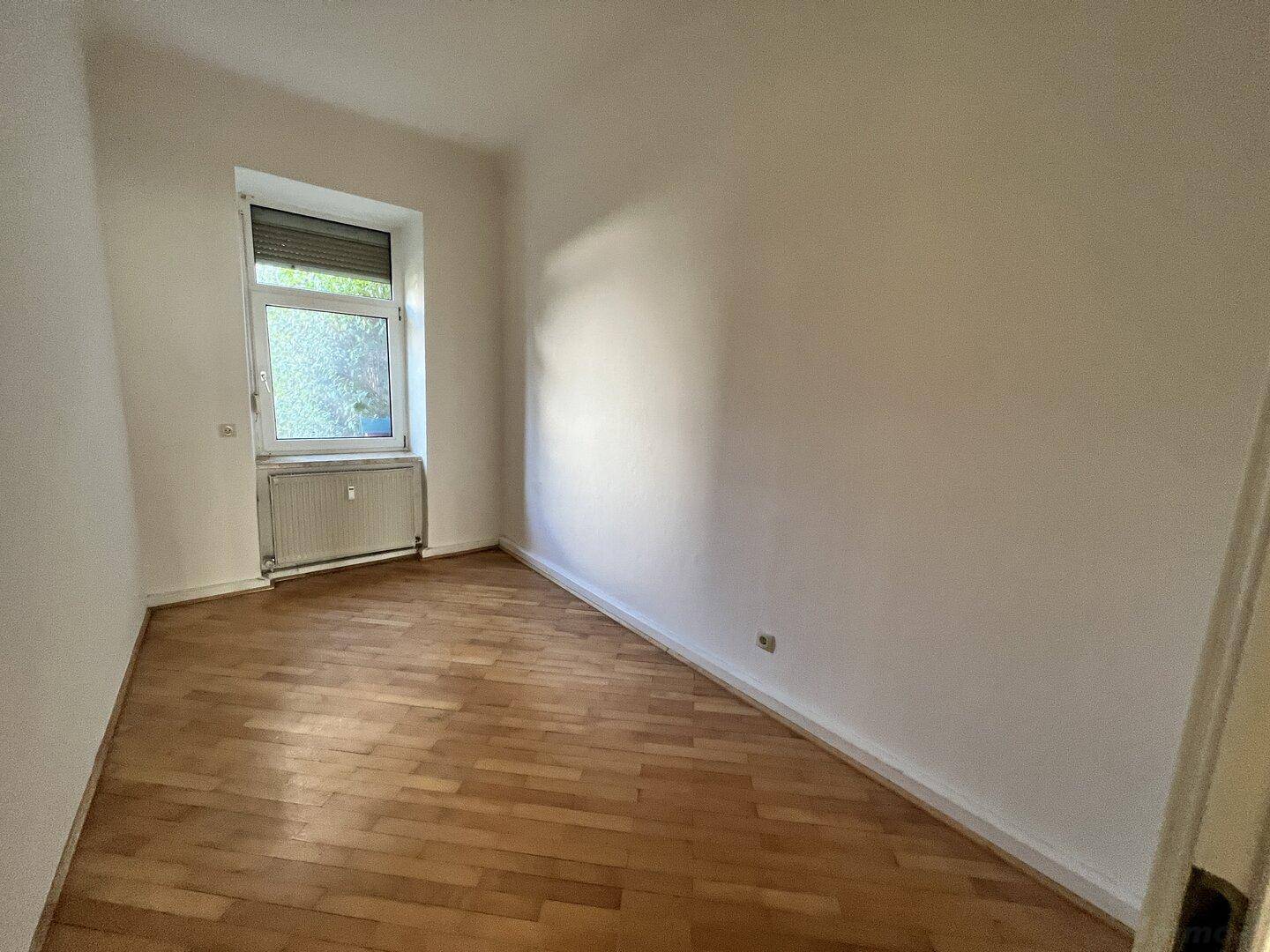 Helles Schlafzimmer mit Holzplankenboden, weißen Wänden und einem Fenster mit Rollladen und Heizkörper.