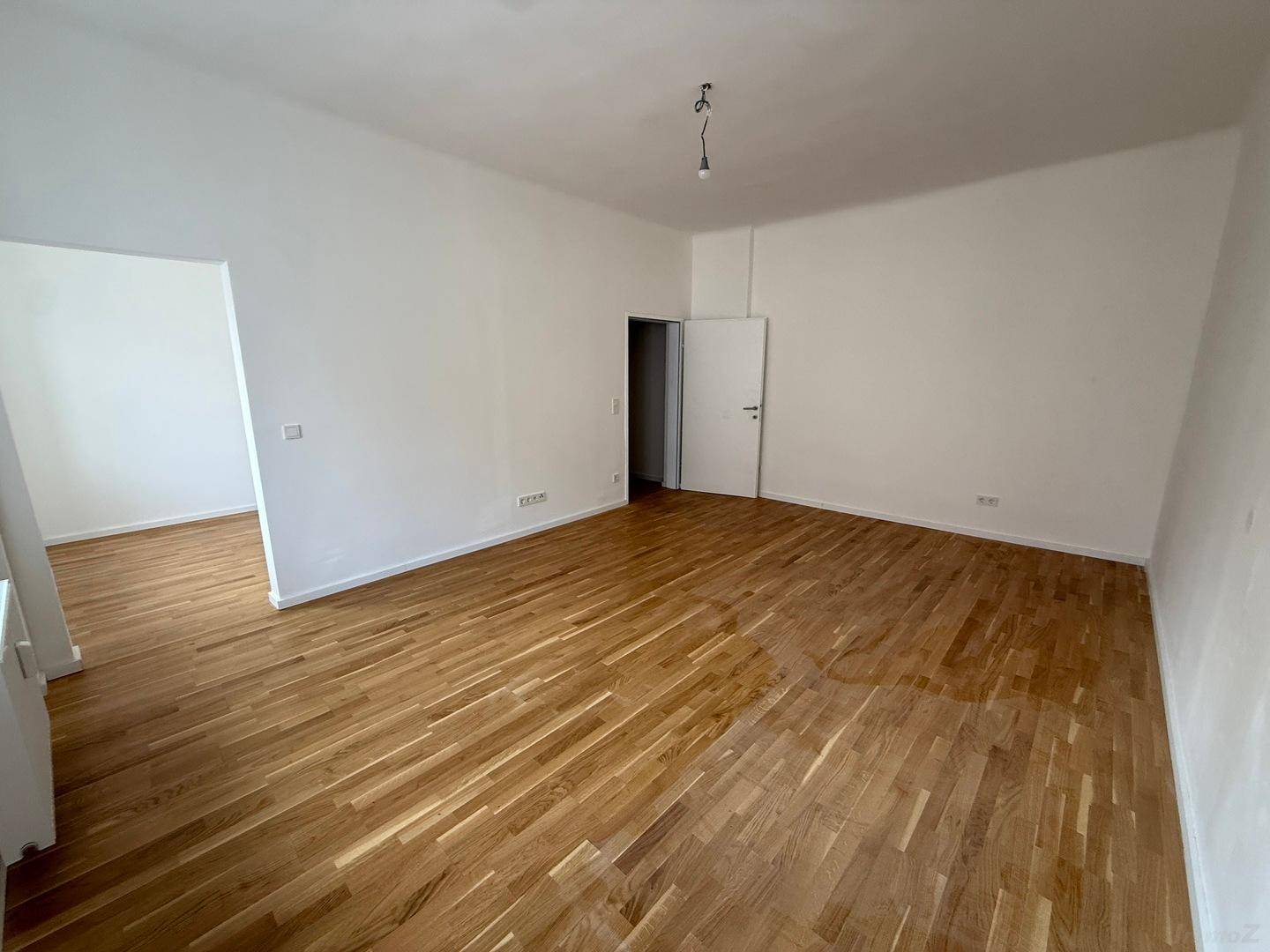 Helles Zimmer mit Holzboden, weiß gestrichenen Wänden und offenem Übergang zu einem Nebenraum.