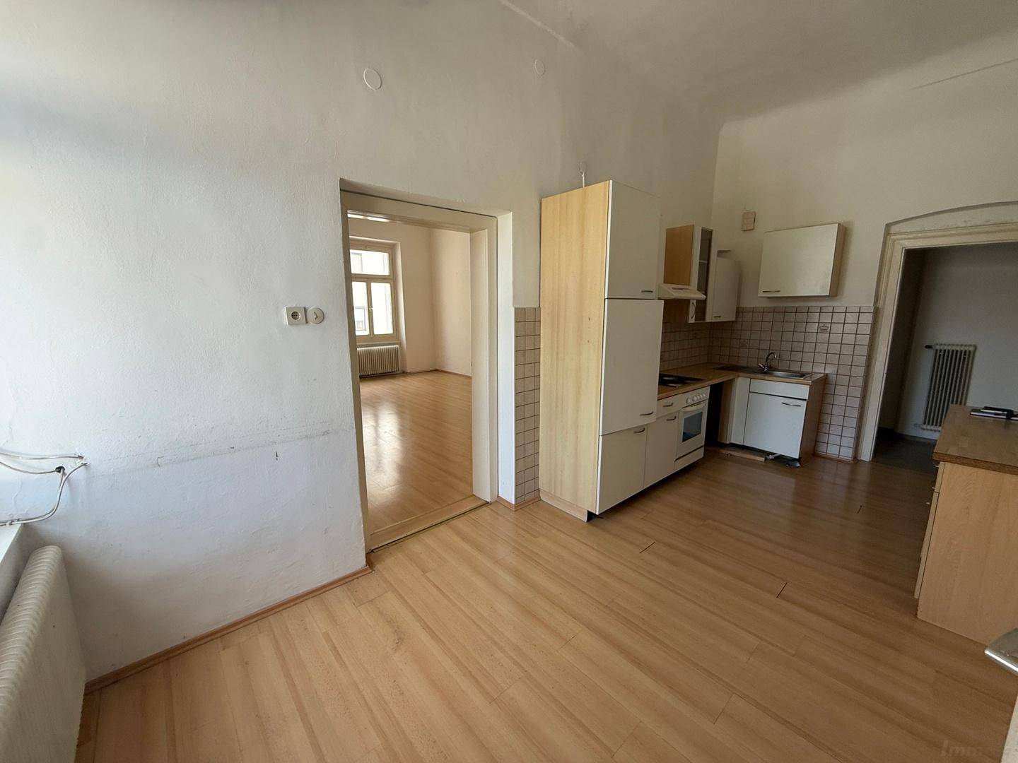 Küche mit hellem Holzboden und Blick durch eine offene Tür in ein angrenzendes, leeres Zimmer.