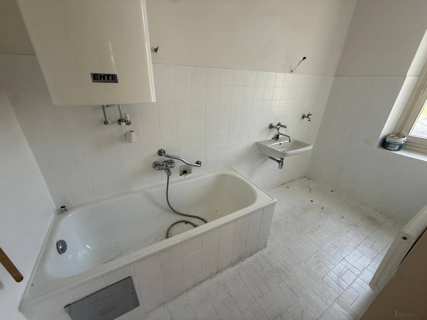Badezimmer mit Badewanne, Waschbecken, Warmwasserboiler und weißen Fliesen, inklusive Fenster.