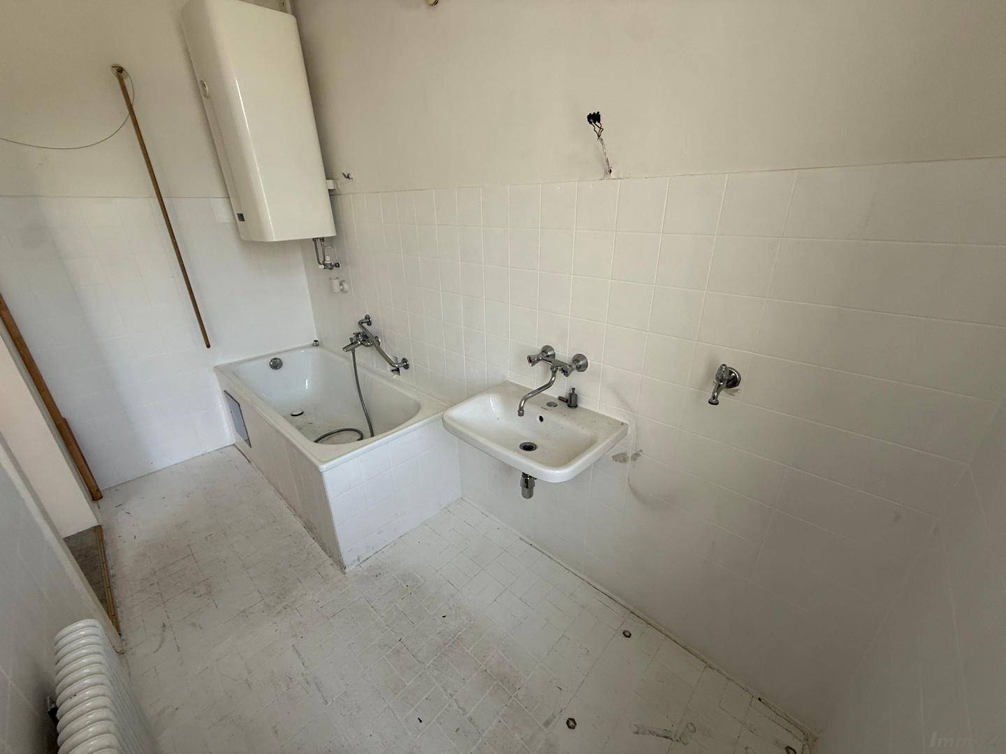 Weiß gefliestes Badezimmer mit Badewanne, Waschbecken und einem Warmwasserboiler an der Wand.