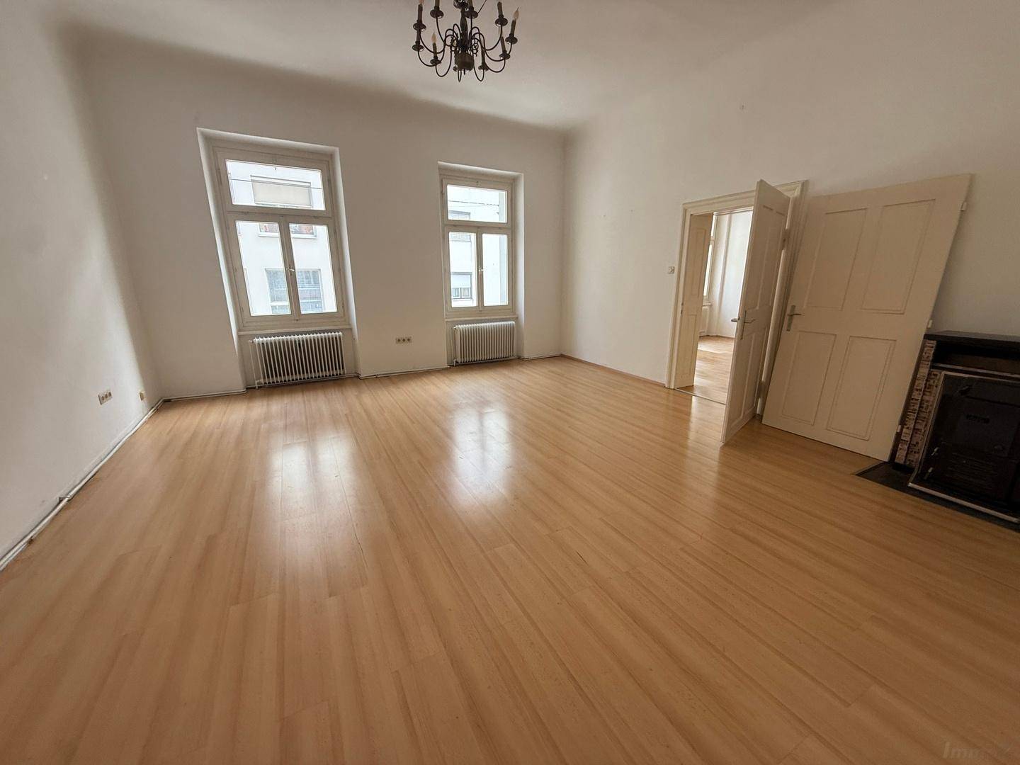 Geräumiges Wohnzimmer mit Holzboden, zwei Fenstern, Kronleuchter und einem Kamin auf der rechten Seite.
