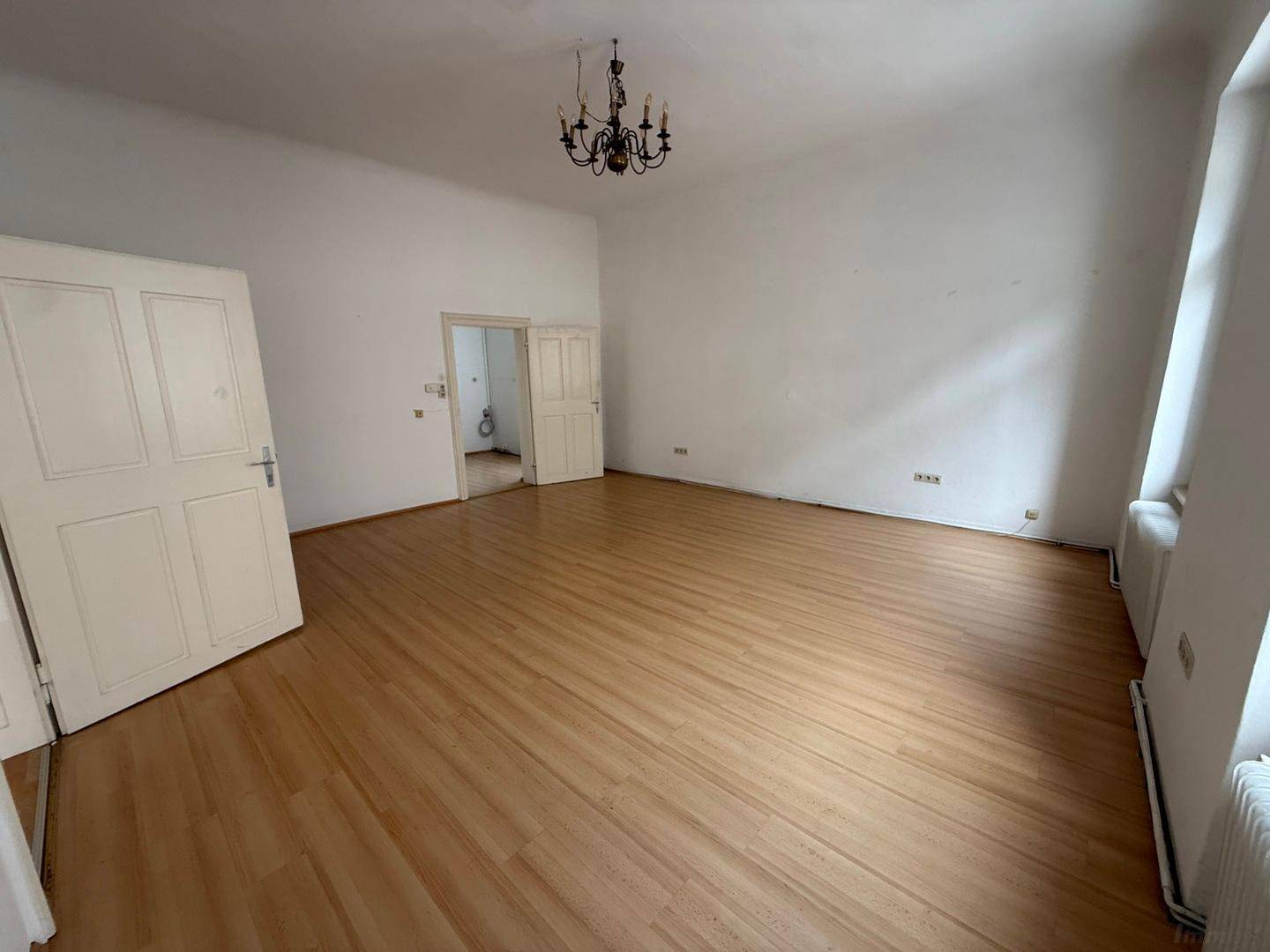 Helles, leeres Zimmer mit Holzboden, weißen Wänden und einer großen, geöffneten Tür.