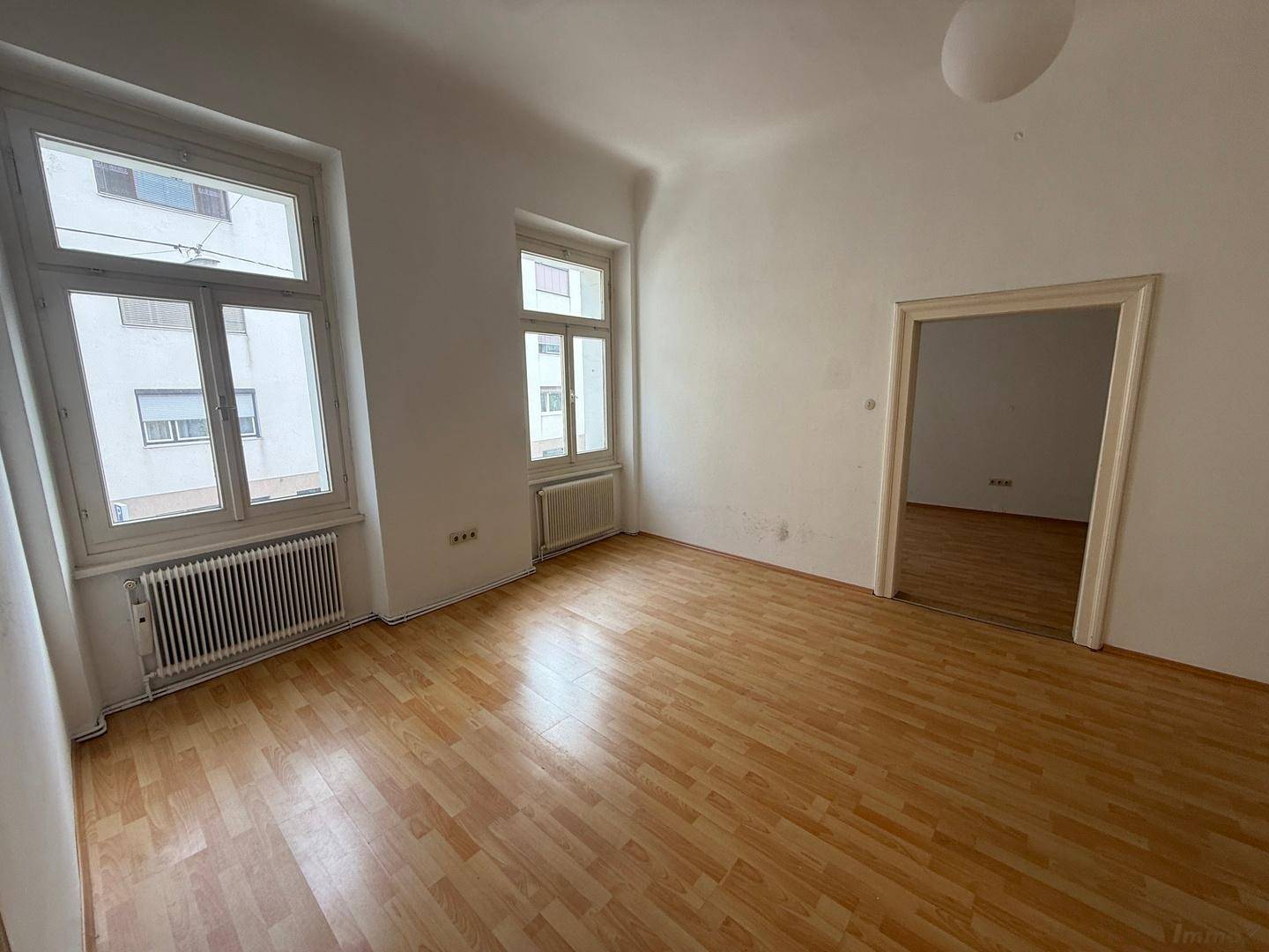 Leeres Zimmer mit hellem Holzboden, zwei Fenstern und einer offenen Tür zu einem weiteren Raum.