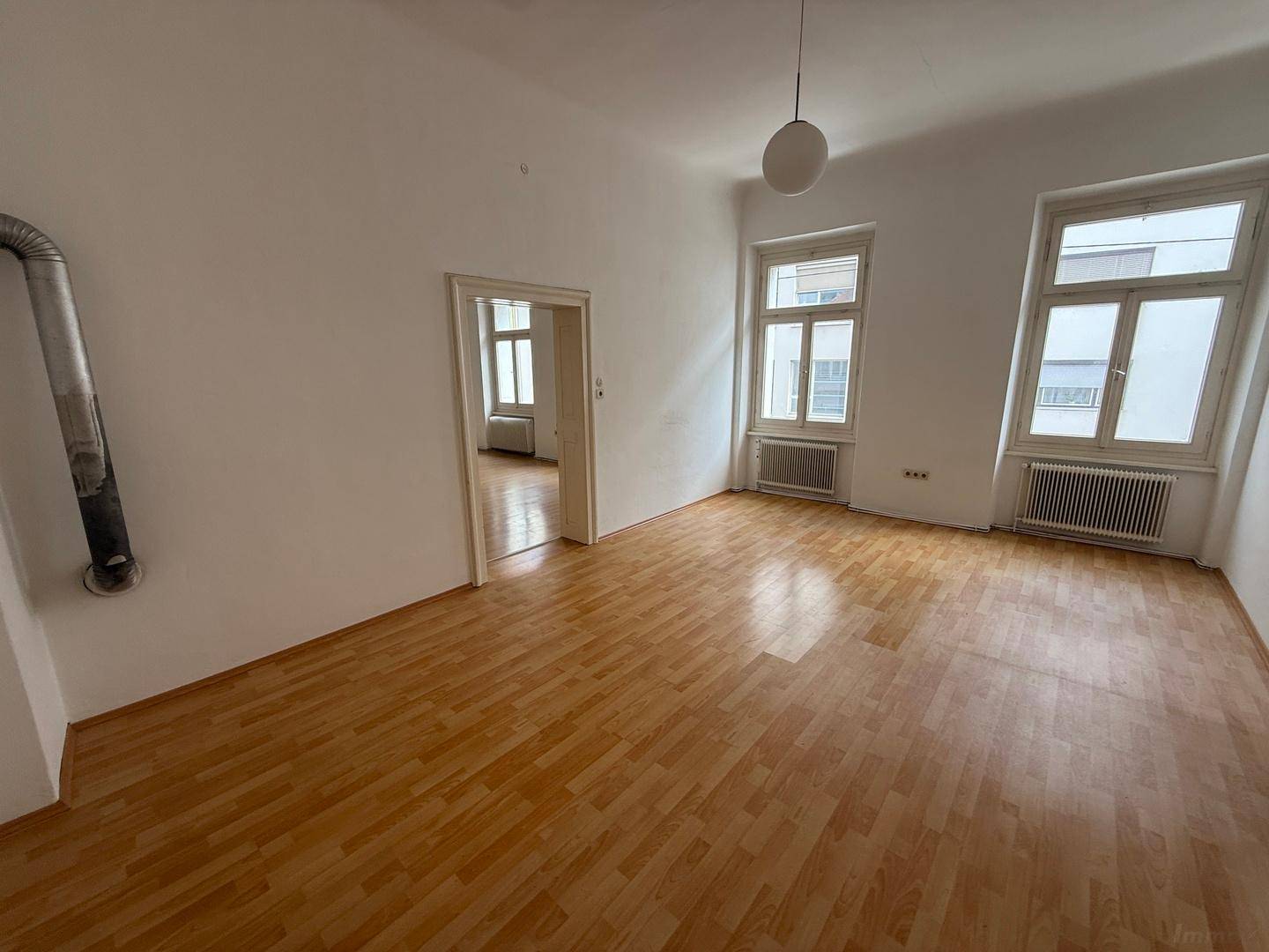 Helles Zimmer mit Holzboden, weißen Wänden, zwei Fenstern und einem sichtbaren Ofenrohr.