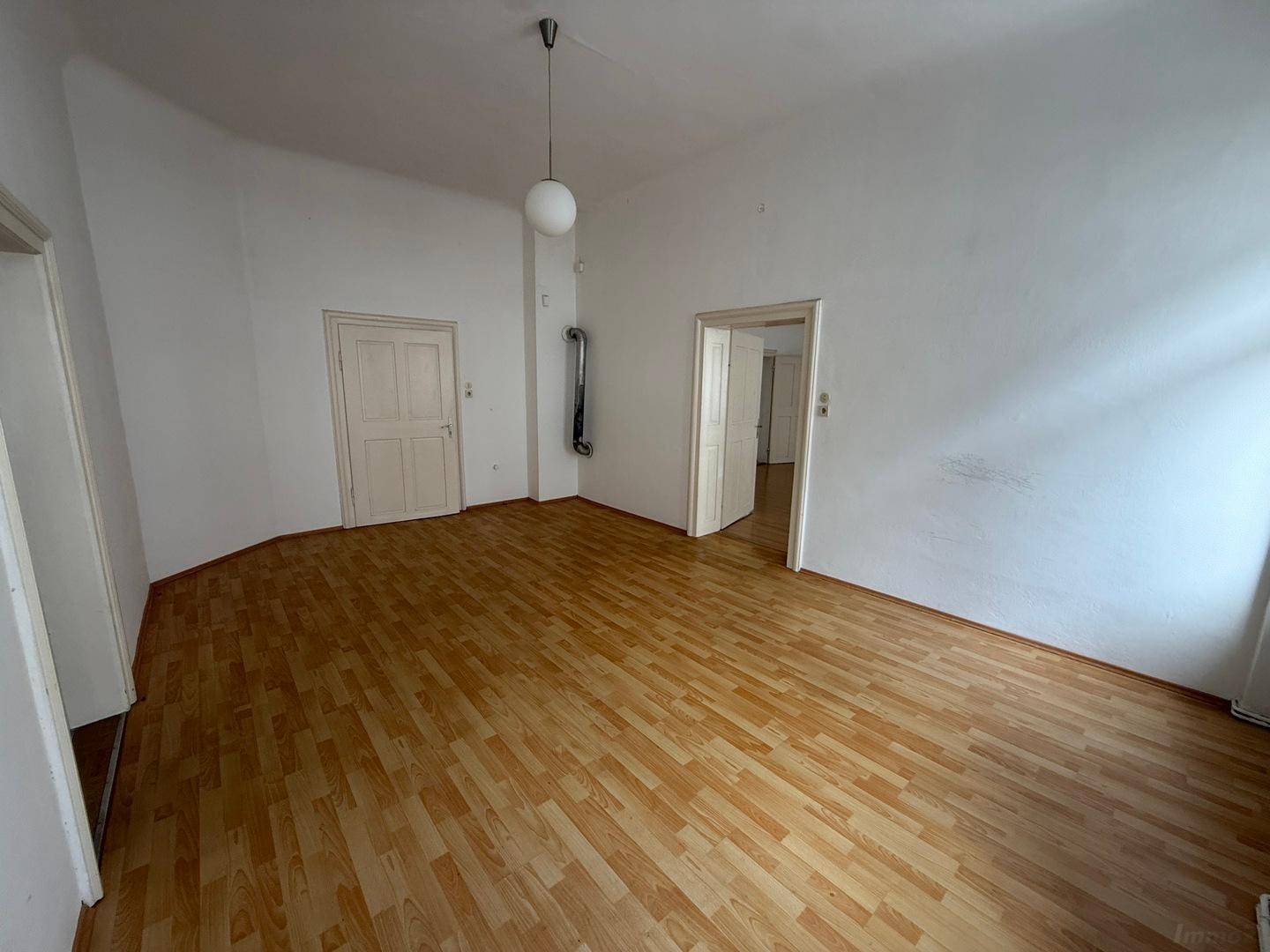 Geräumiges Zimmer mit Holzboden, weißen Wänden und mehreren Türen, inklusive Ofenrohr.