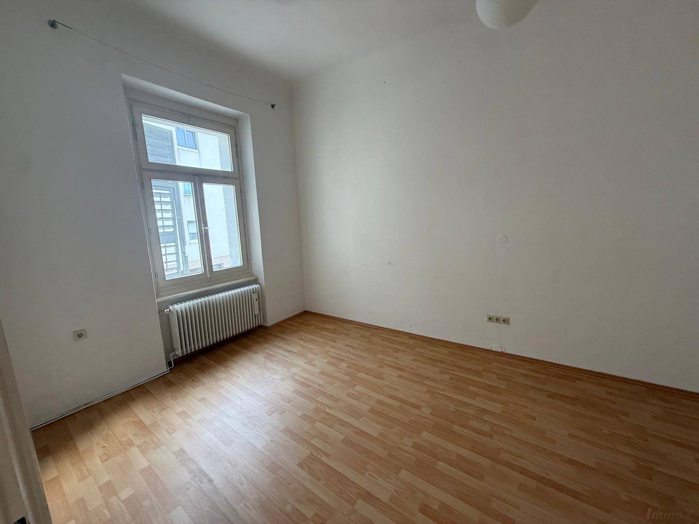 Helles, leeres Schlafzimmer mit Holzboden, weißen Wänden und einem großen Fenster.