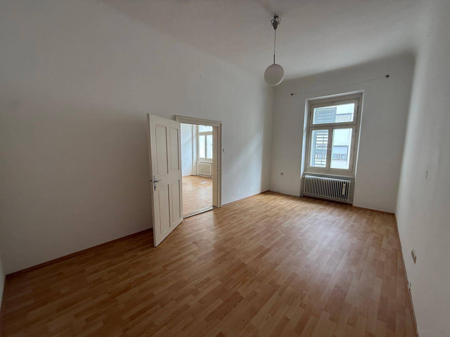 Leeres Schlafzimmer mit Holzboden, weißen Wänden, einem Fenster und einer offenen Tür.