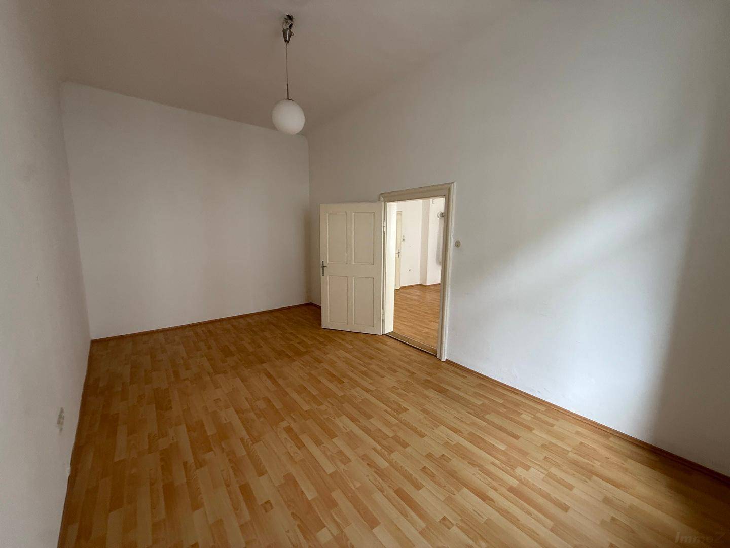 Helles, leeres Schlafzimmer mit Holzboden, weißen Wänden und einer geöffneten Tür.