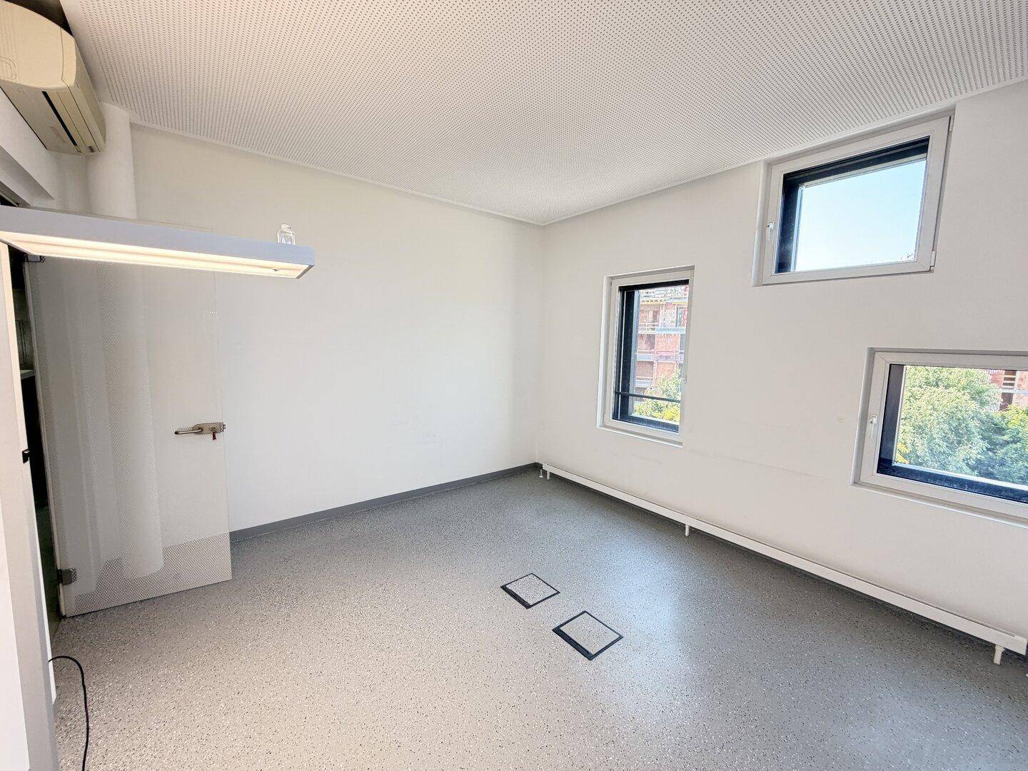 Geräumiger Büroraum mit mehreren Fenstern, Vinylboden und einer Klimaanlage.