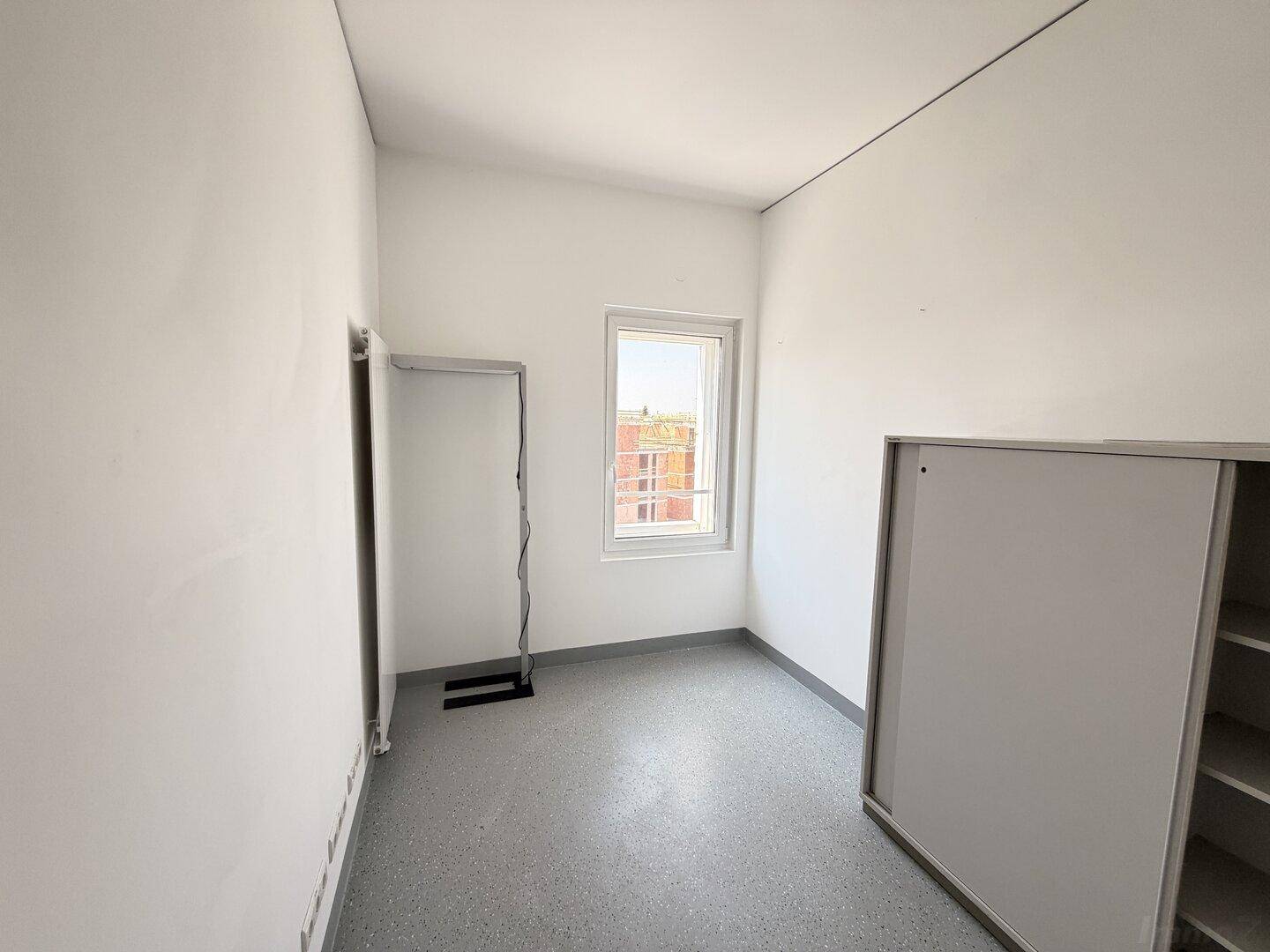 Einfacher Büroraum mit weiß gestrichenen Wänden, Vinylboden und einem Fenster.