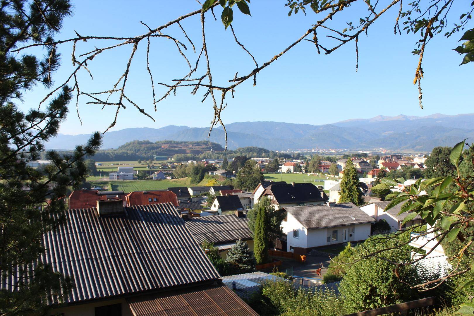 Panoramablick über ein Dorf mit Häusern und Bergen im Hintergrund, umrahmt von Ästen.