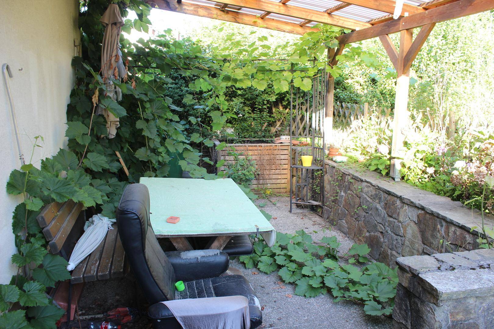 Überdachter Sitzbereich im Freien mit Tisch und Bank, umgeben von üppigem Grün und Weinreben.
