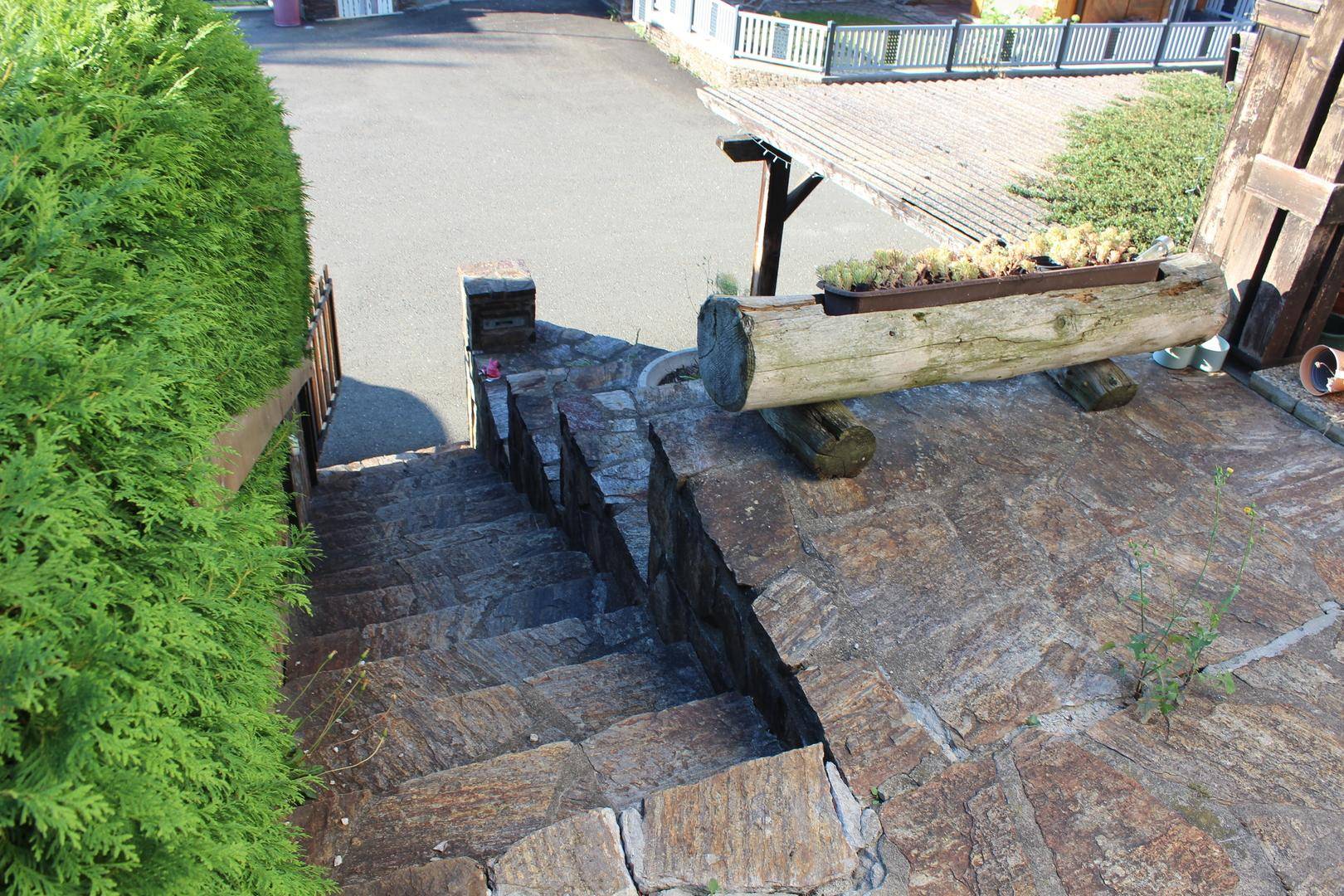 Steintreppe führt hinunter zum Eingangsbereich, umgeben von grünen Hecken und einem Holzbrunnen.