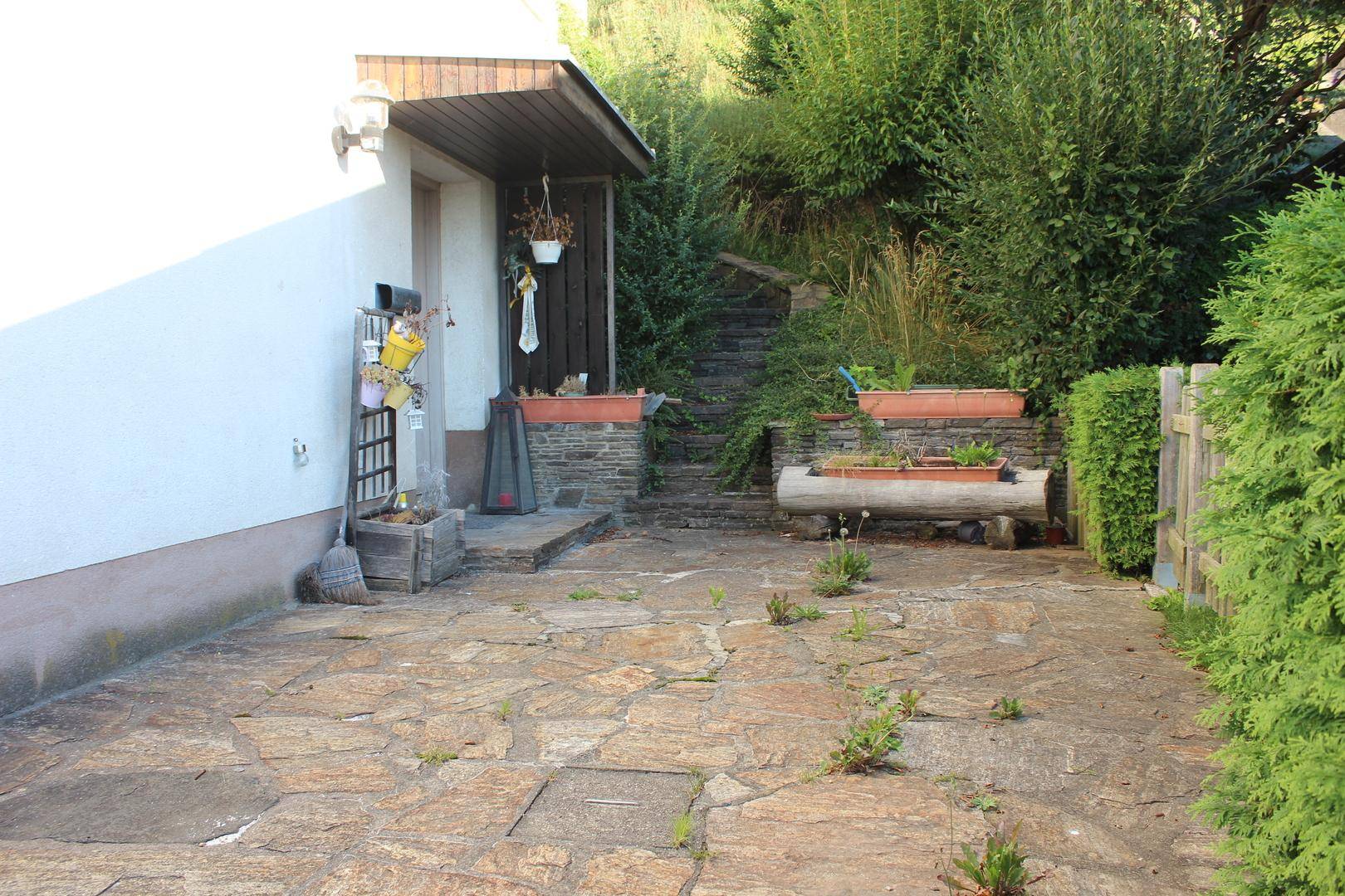 Gepflasterter Terrassenbereich neben dem Hauseingang mit Zugang zu einer Steintreppe und viel Grün.