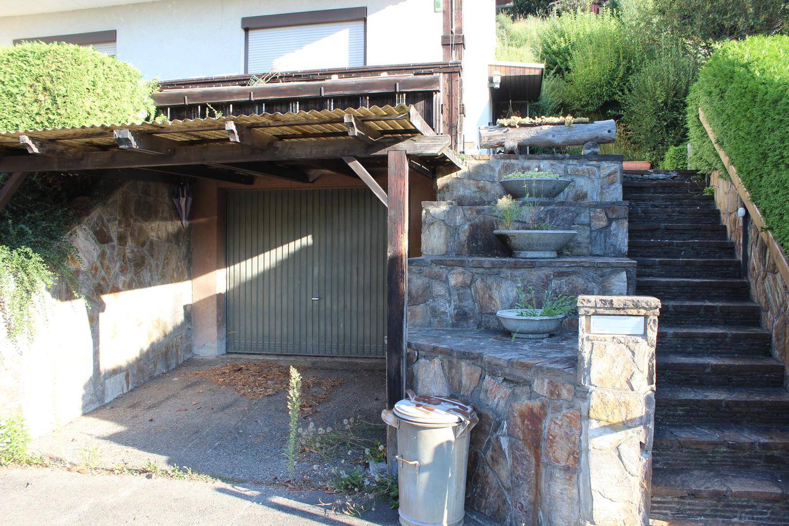 Eingangsbereich mit Carport, Steintreppe und dekorativen Pflanzgefäßen, umgeben von Natursteinmauern.