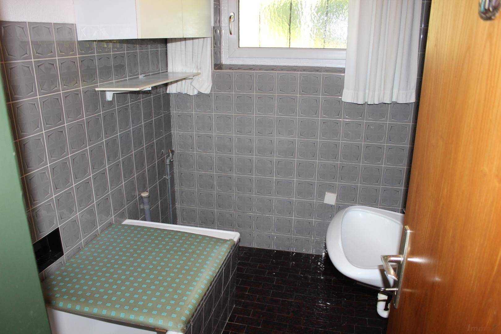 Kleines WC mit grauen Fliesen, Bidet und einem Fenster für natürliche Belüftung.