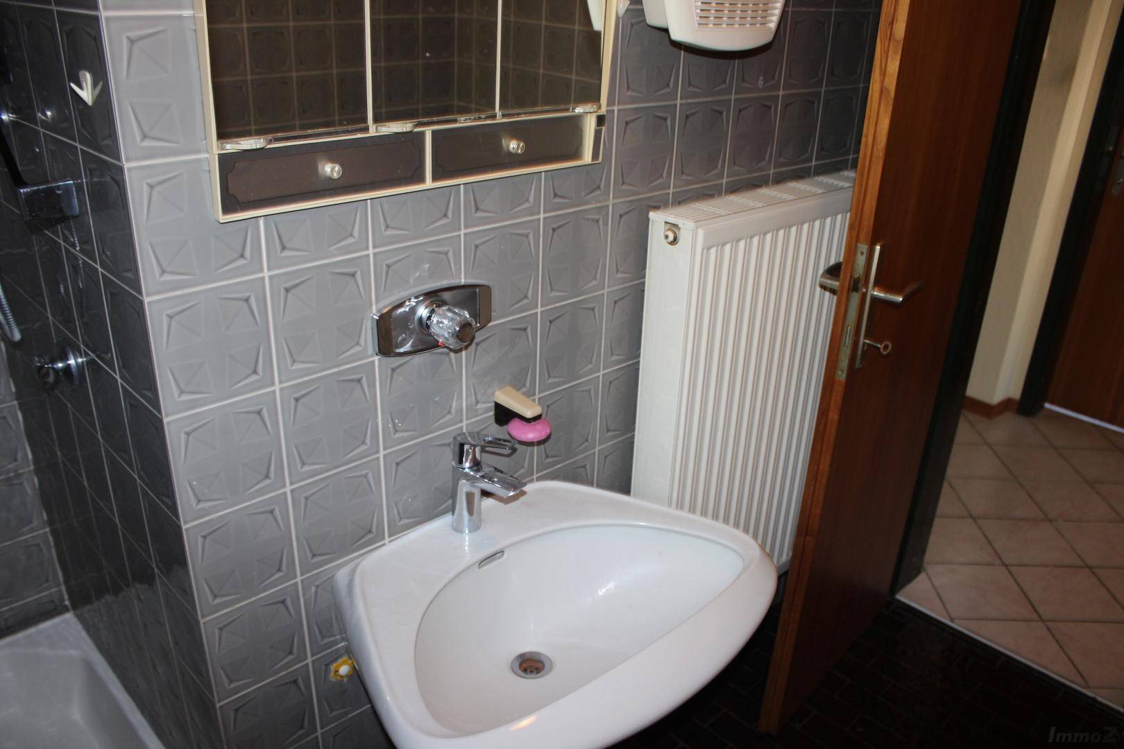 Badezimmer mit grauen Fliesen, einem Waschbecken und einem Heizkörper, im Stil der 80er Jahre.