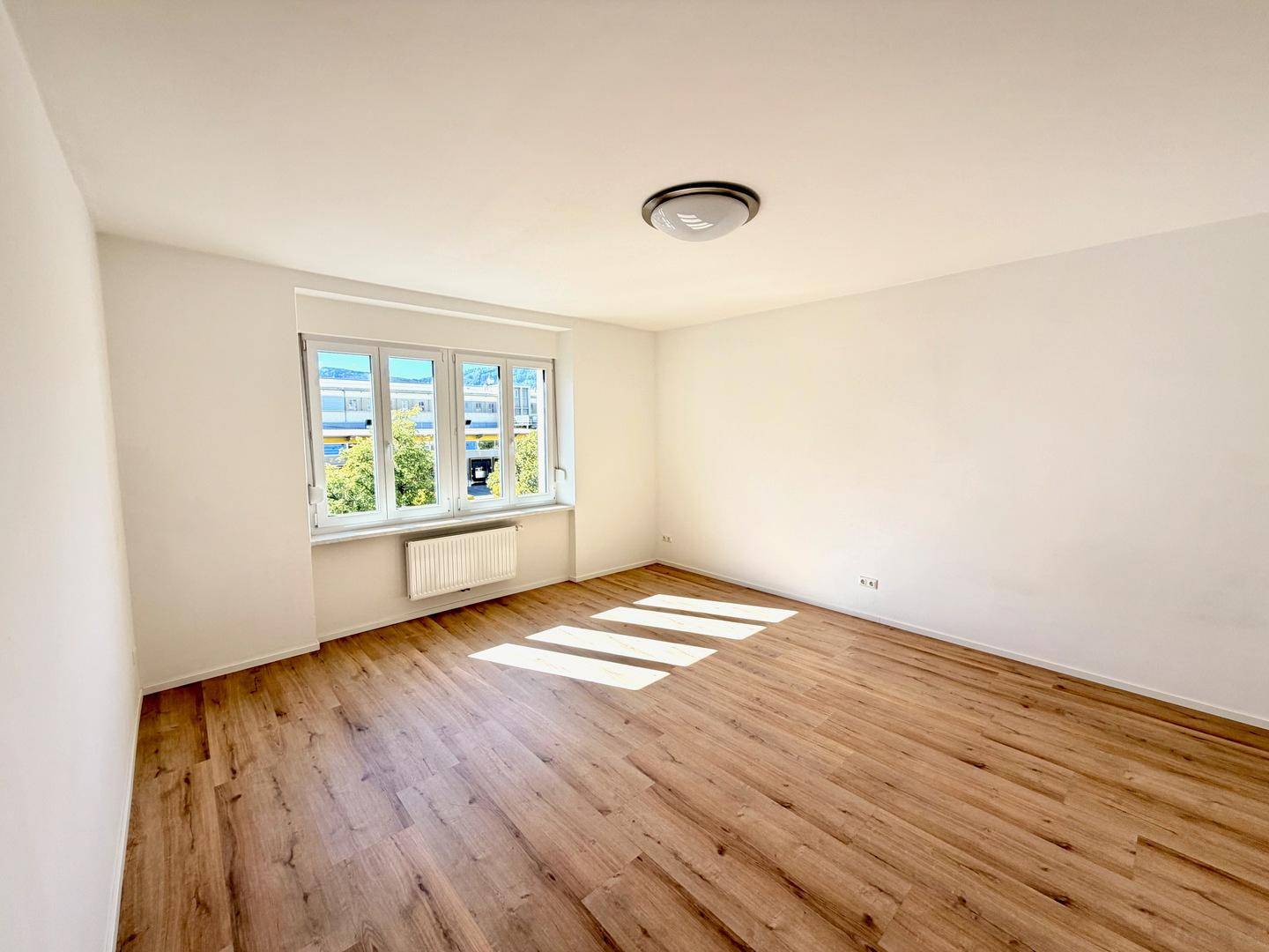 Helles Zimmer mit Laminatboden, weißen Wänden und Blick ins Grüne durch das Fenster.