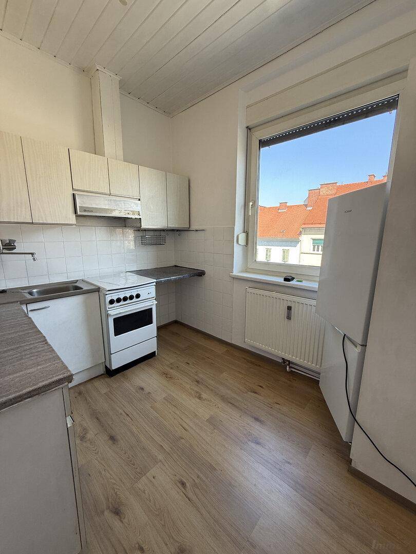 Funktionale Küche mit älteren Einbauschränken, Herd, Spüle und Kühlschrank, mit Blick nach draußen.